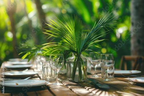 Outdoor dining scene with wooden table set in rich tropical foliage. Glassware, plant vase adorn table. Warm sunlight highlights textures, colors. Perfect for festive banquet or special occasion. photo