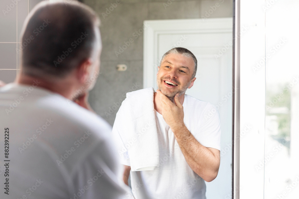 Fototapeta premium Middle aged man touching unshaven chin before shaving and smiling to his reflection in mirror standing in bathroom. Facial hair removal and skincare