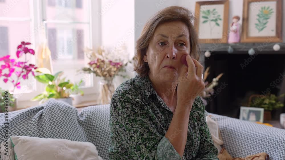 Older woman applying makeup while sitting on a cozy couch in a well-decorated living room, surrounded by plants and framed artwork, creating a warm and personal atmosphere Older woman applying makeup while sitting on a cozy couch in a well-decorated living room, surrounded by plants and framed artwork, creating a warm and personal atmosphere