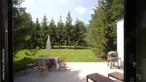Serene outdoor patio with wooden chairs, table and green lawn with pine trees in the background under clear sky. Serene summer environment featuring furniture for family recreation and relaxation
