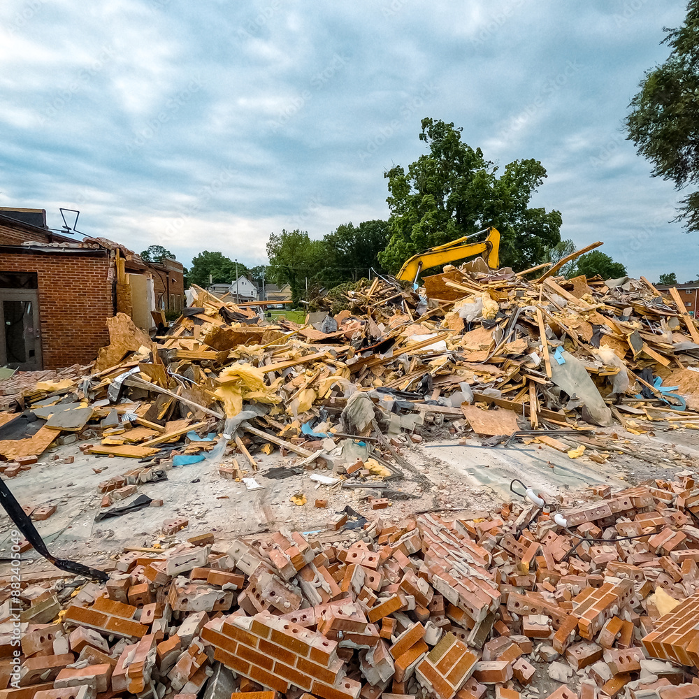 Fototapeta premium A large brick building has just been demolished. A pile of red bricks, wood insulation and debris are stacked in a pile. Bulldozer in background of view. Large demolition construction site.