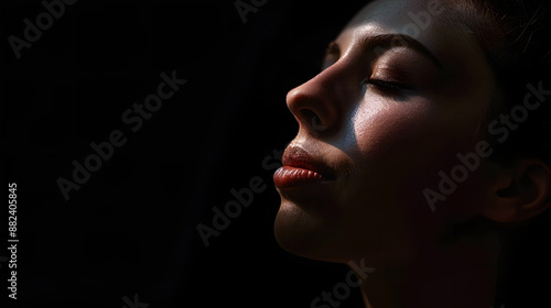 A close up shot of a womans face with eyes closed, lit from the side, with a dark background. She is turned slightly away from the camera. Her lips are closed and she appears to be relaxed.