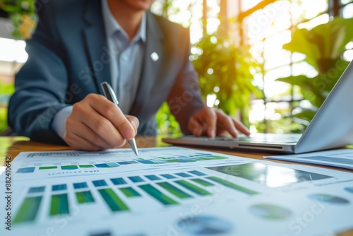 Business person analyzing financial charts and working on a laptop at a desk with plants in the background