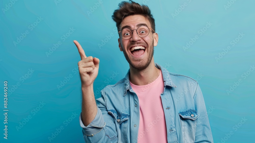 Naklejka premium A young man with glasses, dressed in a denim jacket and pink t-shirt, pointing up excitedly against a blue backdrop, exemplifying enthusiasm and youthful energy.