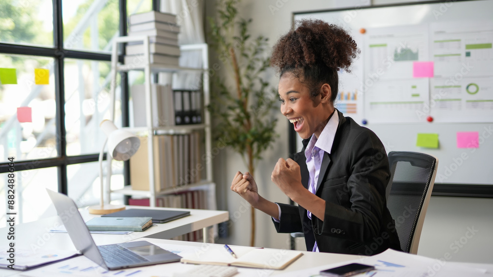 Young professional woman is raising her fists in the air while sitting at her desk, excited about the good news she received on her laptop Young professional woman is raising her fists in the air while sitting at her desk, excited about the good news she received on her laptop