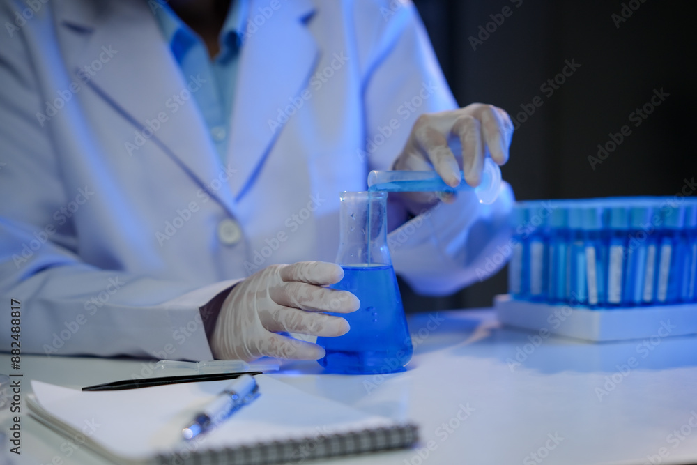 Scientist wearing a lab coat and gloves is carefully pouring a blue solution from a test tube into a beaker, surrounded by scientific equipment in a laboratory setting Scientist wearing a lab coat and gloves is carefully pouring a blue solution from a test tube into a beaker, surrounded by scientific equipment in a laboratory setting
