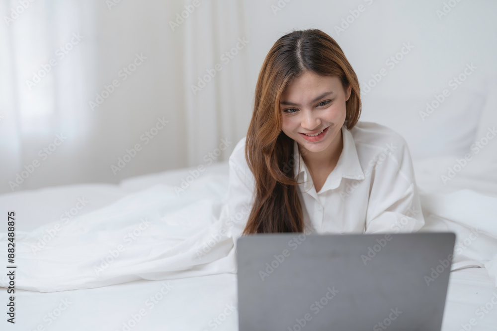 Asian woman happily using her laptop in bed, browsing the internet, smiling and relaxed. Enjoying the convenience of technology at home Asian woman happily using her laptop in bed, browsing the internet, smiling and relaxed. Enjoying the convenience of technology at home