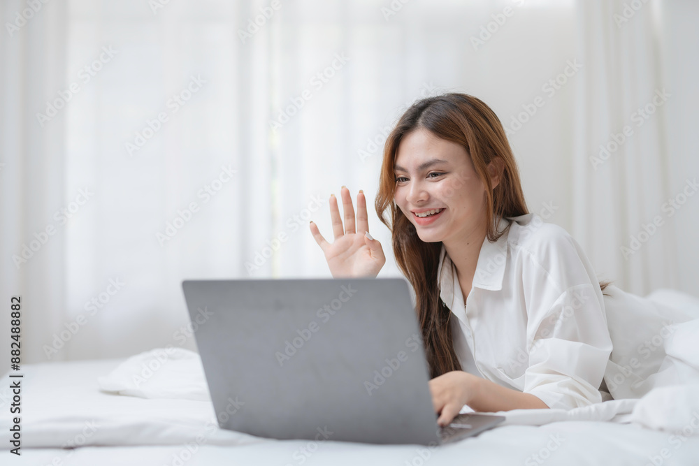 Young woman in bed, working on her laptop and waving during a video call, embodying the modern remote work lifestyle with a smile Young woman in bed, working on her laptop and waving during a video call, embodying the modern remote work lifestyle with a smile