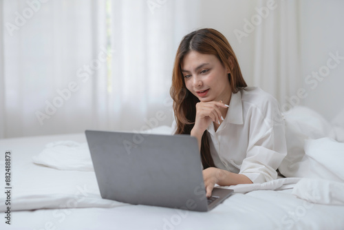 Young woman happily browsing the internet on her laptop in bed, enjoying a cozy morning in her modern bedroom, balancing work and relaxation at home