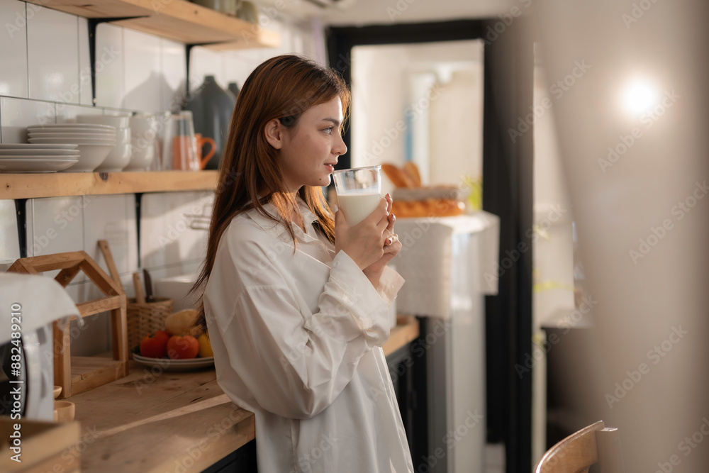 Young woman is standing in her kitchen, holding a glass of milk. She is wearing a white shirt and has long, brown hair Young woman is standing in her kitchen, holding a glass of milk. She is wearing a white shirt and has long, brown hair