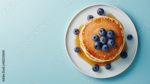 A flat lay of a white plate with a stack of pancakes topped with blueberries and maple syrup, on a light blue background