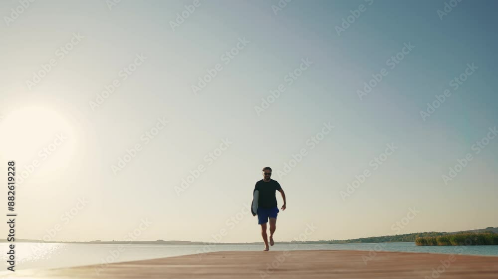A male surfer runs along a pier on a lake against the sun, holding a water board. A male surfer walks along the pier with a small surfboard in his hands. Summer water sports A male surfer runs along a pier on a lake against the sun, holding a water board. A male surfer walks along the pier with a small surfboard in his hands. Summer water sports