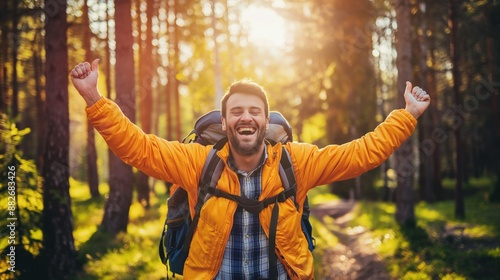 A man in a yellow jacket with a backpack smiles and raises his arms in victory, having reached the summit of a trail. Sunlight streams through the trees of the forest