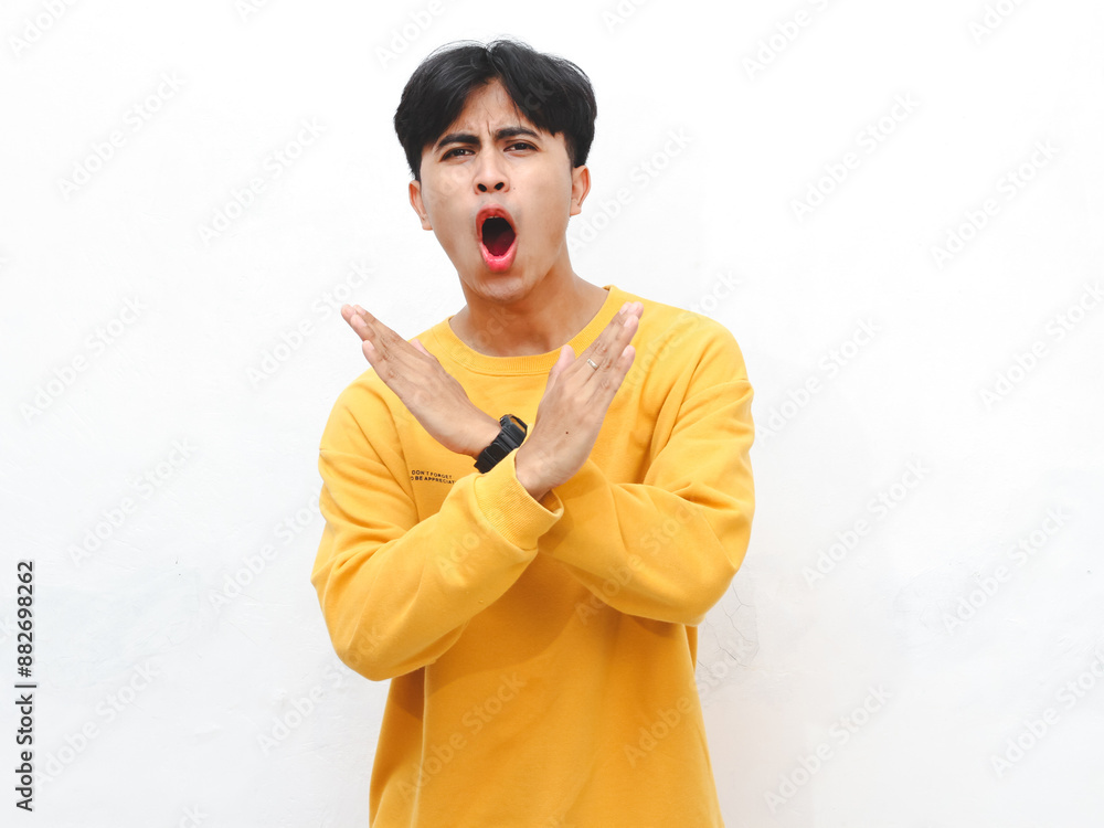 Φωτογραφία Portrait of an Asian young adult man wearing a yellow sweater, showing an angry expression and making a stop gesture with his hand, isolated on a white background