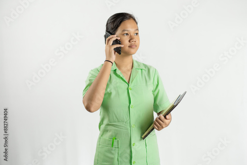 Young Asian female nurse carrying clipboard while making voice call on cell phone, concept of health treatment, working wholeheartedly and dedicatedly, isolated white background.
 photo