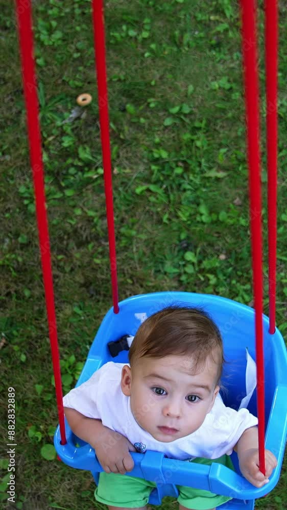 Curious small kid swaying in the swing. Baby looks up at camera. View from above. Green grass at backdrop. Vertical video Curious small kid swaying in the swing. Baby looks up at camera. View from above. Green grass at backdrop. Vertical video