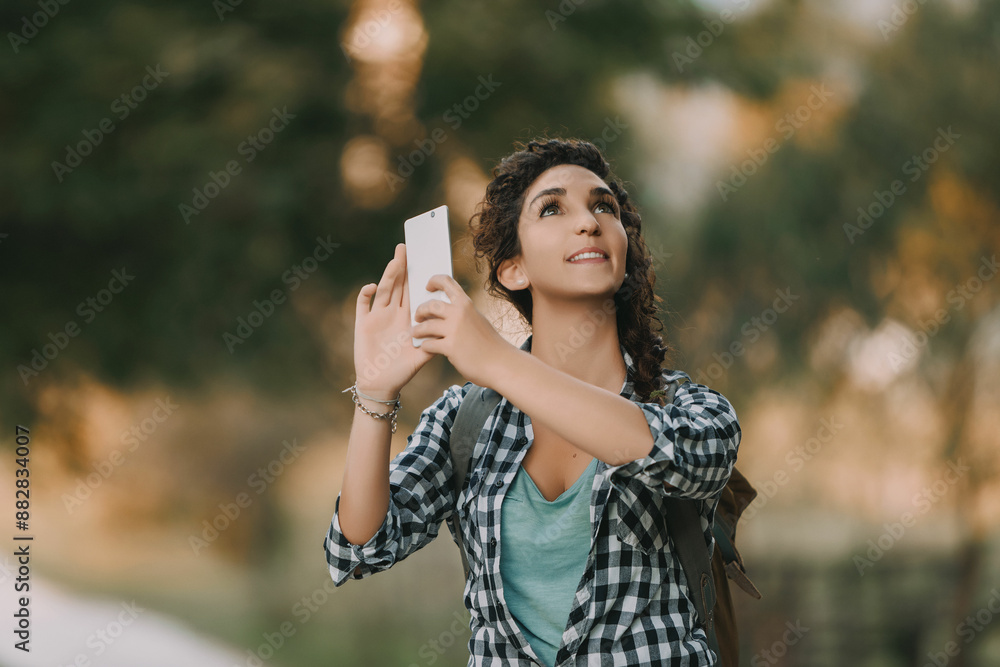Naklejka premium Young woman with curly hair in a braid, dressed in a checkered shirt and green top, holds a smartphone. Her joyful expression and upward gaze indicate she is taking a photo outdoors