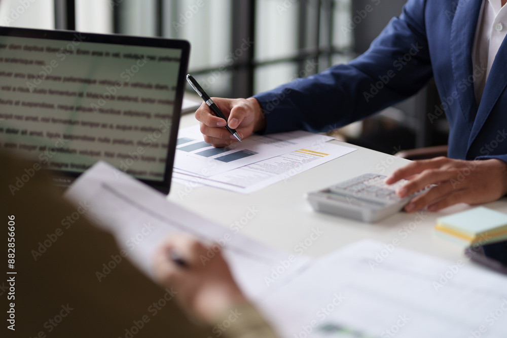 professional setting featuring two individuals working at a table. They are engaged in reviewing documents and using a calculator. Laptops are open on the table. professional setting featuring two individuals working at a table. They are engaged in reviewing documents and using a calculator. Laptops are open on the table.
