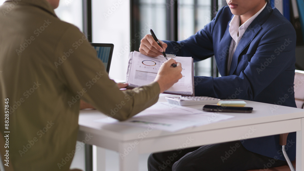 Two individuals are engaged in a business discussion, focusing on a document with graphs and charts. One person is pointing with a pen while the other is holding a clipboard Two individuals are engaged in a business discussion, focusing on a document with graphs and charts. One person is pointing with a pen while the other is holding a clipboard