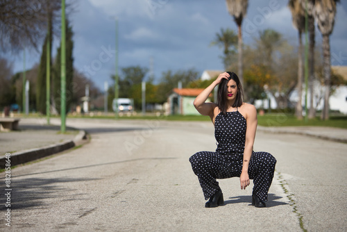 A young, beautiful woman in a black dress with white polka dots, crouched down, sad and alone, in the middle of a road. Concept loneliness, sorrow, beauty, fashion.