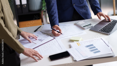 Two individuals standing at a desk, reviewing and discussing documents. A laptop, calculator, smartphone, and various papers with charts and graphs are spread out on the desk