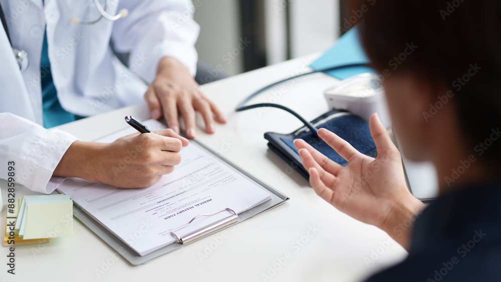 A doctor and patient during a consultation. The doctor is writing on a clipboard while the patient is explaining their symptoms. Medical bottles and a blood pressure monitor are visible on the table A doctor and patient during a consultation. The doctor is writing on a clipboard while the patient is explaining their symptoms. Medical bottles and a blood pressure monitor are visible on the table