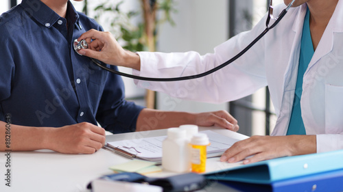 A doctor using a stethoscope to listen to a patient's heartbeat. The patient is wearing a blue shirt and is seated. Medical documents and bottles are visible on the table