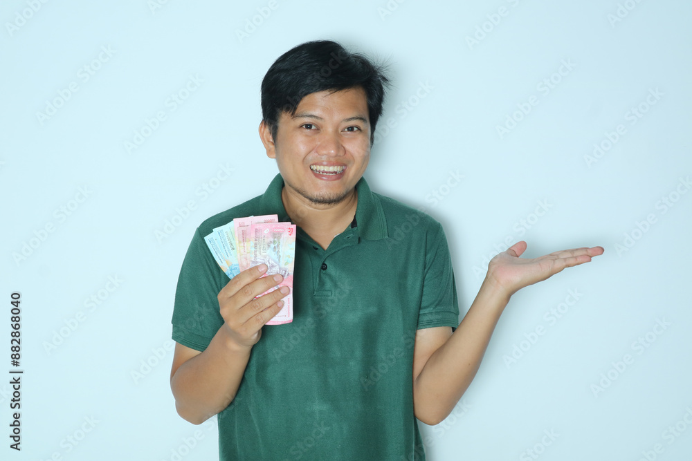 Young Asian man showing smiling happy while holding money with his open left hand while looking to camera. Wearing a green t-shirt Young Asian man showing smiling happy while holding money with his open left hand while looking to camera. Wearing a green t-shirt