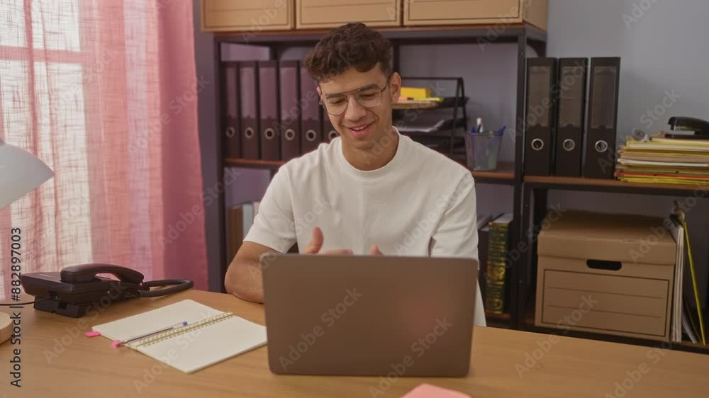 Young hispanic man working on a laptop in an office, smiling and making a celebratory gesture, surrounded by office supplies and shelves with binders and documents. Young hispanic man working on a laptop in an office, smiling and making a celebratory gesture, surrounded by office supplies and shelves with binders and documents.