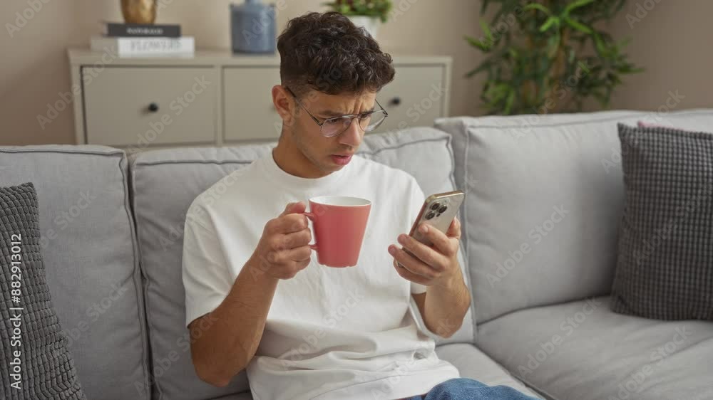 A young hispanic man in glasses drinks coffee while looking at his phone, sitting on a couch in a modern living room with plants and decorations.