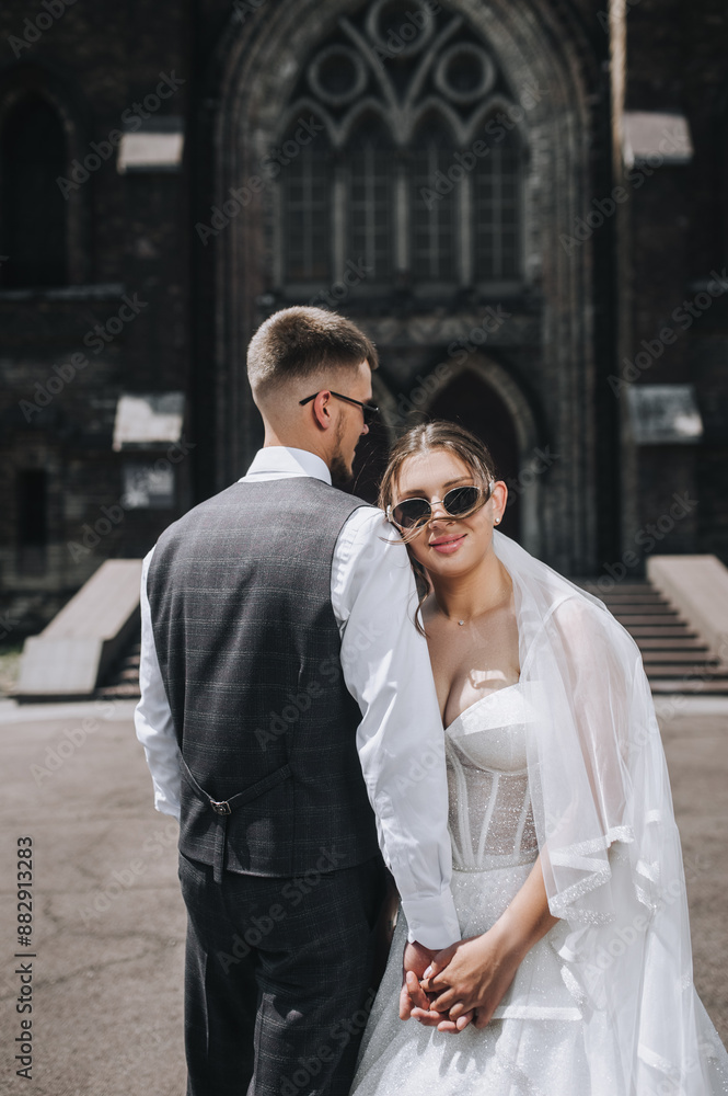 A stylish groom in a gray suit and a beautiful bride in a white dress and sunglasses are standing and hugging against the backdrop of a Christian church. Wedding photography, portrait of newlyweds. A stylish groom in a gray suit and a beautiful bride in a white dress and sunglasses are standing and hugging against the backdrop of a Christian church. Wedding photography, portrait of newlyweds.