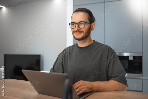 Portrait of young smiling man working home at laptop on kitchen. Business concept. Wearing eyeglasses and sweater.