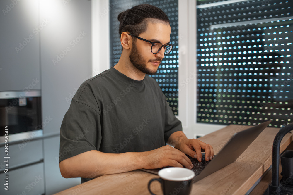 Portrait of young man working home at laptop on kitchen. Business concept. Wearing eyeglasses and sweater. Portrait of young man working home at laptop on kitchen. Business concept. Wearing eyeglasses and sweater.