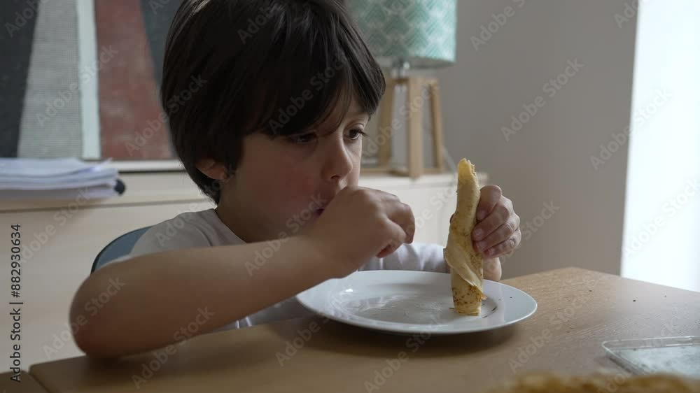 Young boy concentrates on eating a pancake, holding it with both hands and enjoying every bite, creating a heartwarming and candid moment at the dining table Young boy concentrates on eating a pancake, holding it with both hands and enjoying every bite, creating a heartwarming and candid moment at the dining table