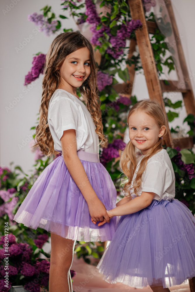 Two young girls, dressed in identical white T-shirts and lilac skirts, stand among the fragrant lilac flowers. Their laughter and smiles radiate the joy of friendship Two young girls, dressed in identical white T-shirts and lilac skirts, stand among the fragrant lilac flowers. Their laughter and smiles radiate the joy of friendship