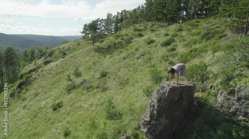 A woman is peacefully practicing a yoga pose on a large rock in a serene natural landscape surrounded by green hills