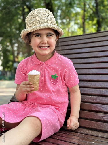 A little girl in a pink dress and hat is eating ice cream in the park in summer. A beautiful brunette girl with ice cream in her hand is sitting on a bench in the park. 