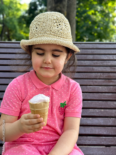 A little girl in a pink dress and hat is eating ice cream in the park in summer. A beautiful brunette girl with ice cream in her hand is sitting on a bench in the park.  