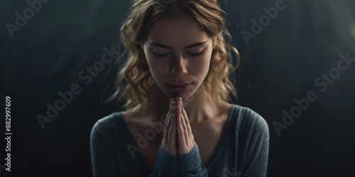 Young woman with blonde hair and blue eyes sits in contemplation. Hands clasped together, she gazes off to side against dark background. Serene expression, peaceful atmosphere.