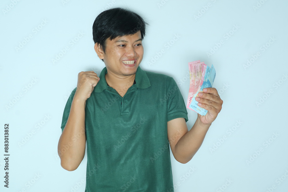 Young Asian man showing excited face expression with fist clenched while holding paper money. Wearing a green t-shirt Young Asian man showing excited face expression with fist clenched while holding paper money. Wearing a green t-shirt