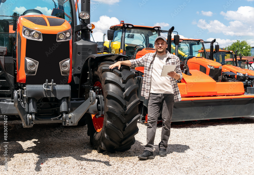 Caucasian adult man with digital tablet in hand stands next to construction or agricultural tractor at a machinery dealer. Ag equipment dealer or supplier. Caucasian adult man with digital tablet in hand stands next to construction or agricultural tractor at a machinery dealer. Ag equipment dealer or supplier.