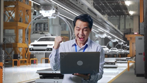 Wallpaper Mural Asian Male Professional Worker Standing With His Laptop In EV Car Factory, High-tech Industrial Facility, He Raises His Fist Up With Screaming Goal Torontodigital.ca