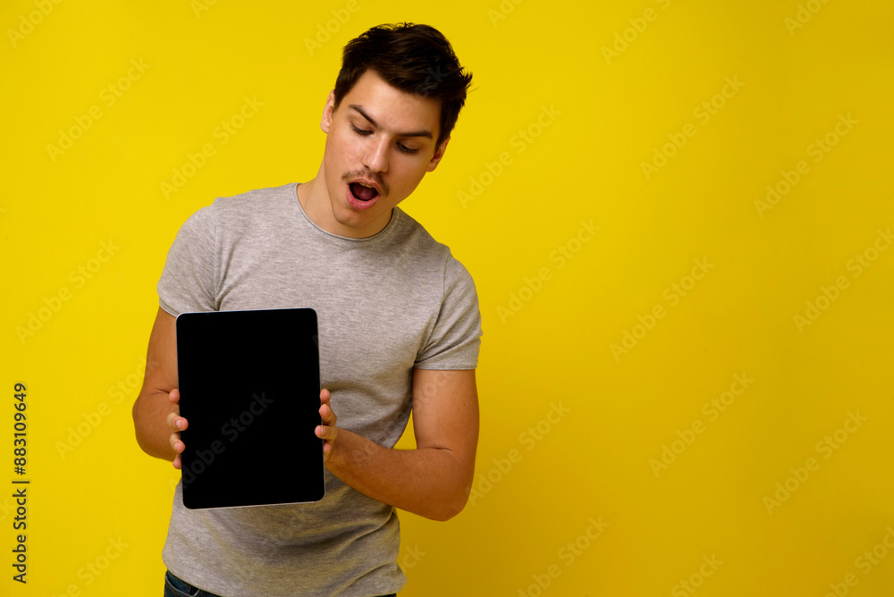 A surprised young man in a gray t-shirt stands against a yellow backdrop, looking at a tablet with amazement and his mouth slightly open A surprised young man in a gray t-shirt stands against a yellow backdrop, looking at a tablet with amazement and his mouth slightly open