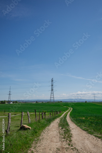 Peaceful summer afternoon, a rural landscape unfolds, featuring a wooden fence alongside a sandy road stretching to the horizon amid green pastures. In the distance, tall power poles are visible