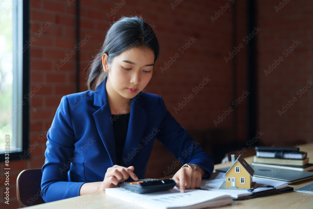 A house seller in a blue suit is working with a calculator at a desk. She is holding a piece of paper and appears focused on her calculations