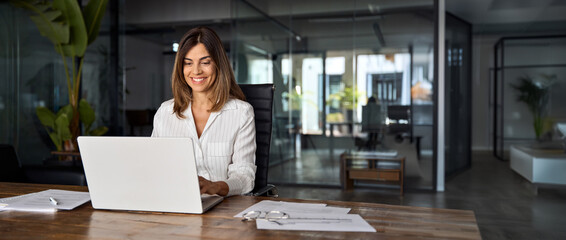 Middle age European business woman CEO using laptop application for work at table workspace in office. Smiling Latin Hispanic mature adult professional businesswoman using pc digital computer. Banner