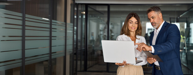 Mature mentor man and European leader woman discussing project on laptop in office. Two diverse partners, colleagues, team of confident professional business people work together. Banner, copy space