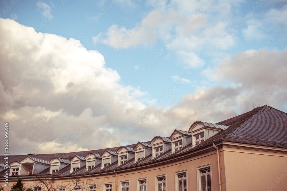 Ein apricot-farbenes, geschwungenes Gebäude mit dunklem Schindel-Spitzdach und vielen weiß-gerahmten (Erker-)Fenstern. Über dem Gebäude sieht man weiße Wolken vor blauem Himmel. Ein apricot-farbenes, geschwungenes Gebäude mit dunklem Schindel-Spitzdach und vielen weiß-gerahmten (Erker-)Fenstern. Über dem Gebäude sieht man weiße Wolken vor blauem Himmel.