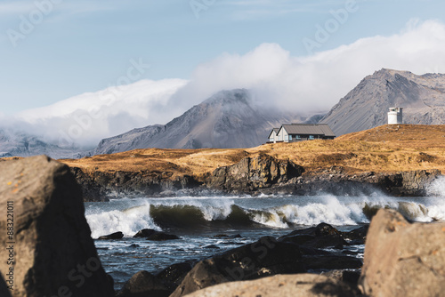 Panoramic view of stormy waves over two iconic buildings to cloud covered mountains on a sunny day at Ytri Tunga Beach. photo