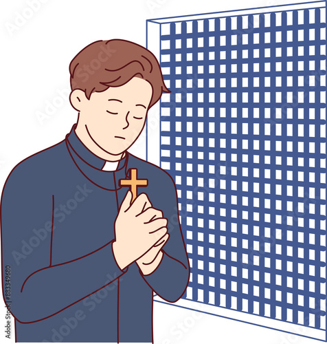 Padre prays and holds christian cross in hands, standing in confession room, asking god to atone for sins of temple visitors. Catholic priest observes ritual of confession and reads prayer